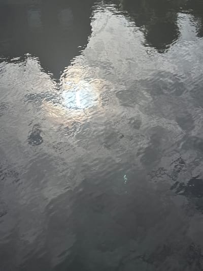 The image shows a close-up, high-angle view of the surface of a body of water, likely a canal or river in Katwijk, Netherlands. The water is dark gray and reflects the sky, which appears overcast with patches of bright sunlight breaking through, creating shimmering highlights on the water's surface. There are faint reflections of what appear to be buildings or structures on the left side of the image, distorted by the ripples of the water. In the lower right quadrant of the image, a small, bright green object floats on the water's surface. The overall impression is one of a natural, calm, and somewhat somber urban waterway scene, typical of Dutch cityscapes.