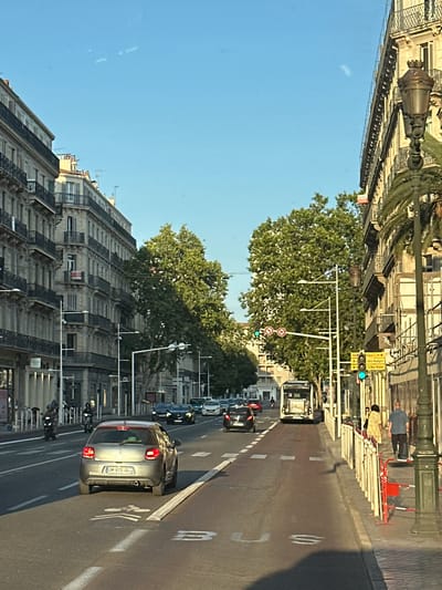 The image shows a wide street in Toulon, France, with ornate, multi-story buildings lining both sides. The street is lined with trees and features a dedicated bike lane marked on the asphalt. Several cars and motorcycles are visible, some in motion and others parked. A bus is also present in the traffic. Pedestrians are seen walking on the sidewalk. Traffic lights and street lamps are positioned along the road. The sky is clear and blue, suggesting a sunny day. The architectural style of the buildings indicates a European city, and the presence of a "Toulon" sign confirms the location.