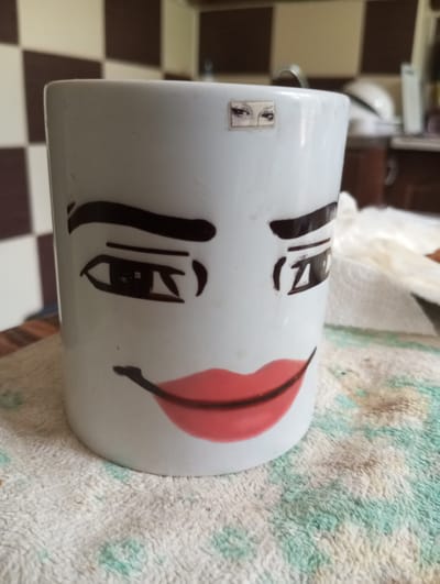 A white ceramic mug with a face drawn on it sits on a patterned, textured surface. The face features drawn-on eyebrows, eyes with visible eyelashes, and red lips. A small, square sticker with two eyes is placed on the upper part of the mug, above the drawn-on face. The background of the image shows a tiled wall with brown and beige squares, and a glimpse of kitchen cabinetry. The scene appears to be indoors. The location in the city of Dymer, Ukraine is not discernible from the image.