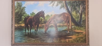 This is a framed painting depicting two horses in a body of water, likely a river or stream, with trees and foliage in the background under a cloudy sky. One horse, darker in color, stands upright and appears to be looking to its left. The second horse, lighter in color, is bent down, drinking from the water. The painting is framed with an ornate, metallic border. The image provided does not contain any information about the city of Chernomorskiy, Russia, or any elements that would indicate this specific location.