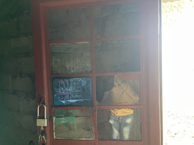 The media file captures a close-up view of a red-framed window or door, likely situated within a building in Nzulo, Democratic Republic of the Congo. To the left, a textured wall constructed from rough, light-colored blocks is visible. Intense daylight floods in from the right side of the frame, causing significant overexposure outside and casting bright reflections on the glass panes.

A silver padlock with keys is attached to a latch on the left side of the red frame, suggesting security. The window or door consists of multiple rectangular glass panes, many of which are reflective or damaged. In the upper left pane, faint, mirrored text is discernible but too blurry and obscured by reflections to be read clearly.

The middle left pane showcases an image, possibly a framed photograph or a screen display, featuring a group of people, likely children or young adults. They are dressed in matching pink, purple, and white attire, gathered in an outdoor setting with green foliage. The glass in the middle right and lower right panes is noticeably cracked and broken. These broken panes distinctly reflect a partial image of a person wearing a yellow t-shirt and blue jeans, presumably the individual taking the photograph.

The scene combines elements of the interior (rough wall, padlock), an external environment (bright daylight), and a personal reflection, layered through the glass. The time of day is clearly bright daylight. No specific activity is depicted other than the act of photography.