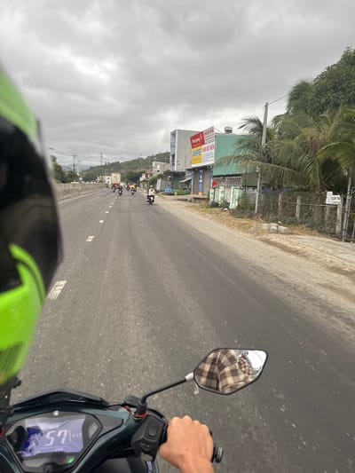 A first-person perspective from a moving motorbike on a straight, paved road. Other motorbikes are visible in the distance. Buildings and vegetation line the road's edge, obscured by overcast skies. The motorbike's instrument panel displays "57". The rearview mirror reflects the rider's plaid-patterned clothing. A hand grips the handlebar. Part of the helmet is visible at the bottom of the frame.