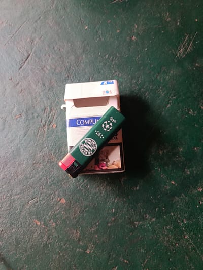 The image shows a close-up, overhead view of a pack of cigarettes and a green lighter resting on a dark green, textured surface, possibly concrete or asphalt. The pack of cigarettes is white with blue and silver accents, featuring text that appears to include "COMPLI" and "6 mg". The lighter is dark green and has a red tip. It is adorned with several symbols, including a soccer ball within a star emblem, the UEFA logo, and a circular crest with a shield and "FC BAYERN MÜNCHEN" text. The lighter is positioned diagonally across the cigarette pack. A portion of the cigarette pack's opening is visible, revealing what looks like the edge of a photograph or graphic containing small stuffed toys. The lighting suggests it is daytime, but no specific weather conditions are discernible. The scene appears to be outdoors in an urban or industrial setting, given the textured ground surface. There are no people visible in the image. The overall impression is a still life arrangement of smoking paraphernalia.