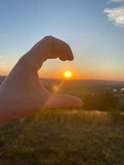A human hand is shown in silhouette, curved to form a C-shape. The sun is positioned within the curve of the hand, appearing as a bright, round orb. The sun's rays create a lens flare. The background depicts a landscape at sunset, with rolling hills and distant trees. The sky is a gradient of blue to orange, with a few scattered clouds. The foreground consists of grassy terrain. The location is stated to be Уязытамак, Russia.