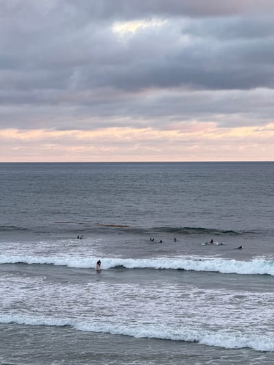 A group of surfers are in the ocean off the coast of Encinitas, California, on a cloudy day. The sky is filled with shades of gray and pink clouds. The ocean is a deep blue-gray, with waves rolling in. Several surfers are visible on their boards, some paddling and others waiting for waves. One surfer is actively riding a wave closer to shore. The scene captures a typical surfing session in Encinitas.