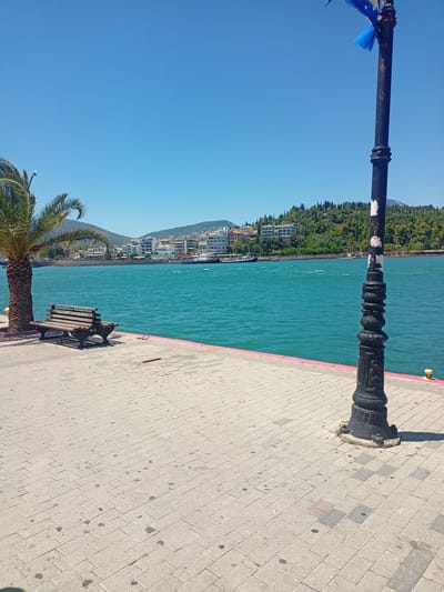 This is a daytime, eye-level, outdoor, full shot of a waterfront promenade in Chalkida, Greece. In the foreground, a paved walkway made of light-colored rectangular stones leads to a bright turquoise body of water. A single wooden bench is positioned near a palm tree on the left side of the frame. A black lamppost with decorative elements and a blue ribbon tied to it stands on the right side of the walkway. The water is choppy, and a white boat with a blue stripe is visible in the middle distance. Across the water, a city skyline with white buildings is nestled against a backdrop of lush green hills. The sky is a clear, bright blue. The scene is devoid of any people.