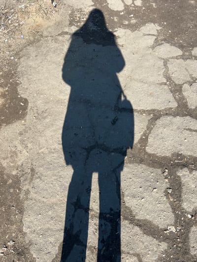 The image displays a full-length shadow of a person cast onto a cracked, uneven ground surface under direct sunlight. The shadow, which belongs to the photographer, depicts an individual wearing what appears to be a long coat or jacket, possibly a head covering, and carrying a bag on their right side.
The ground consists of light grey asphalt or concrete, exhibiting numerous cracks filled with darker dirt and debris. Patches of loose soil, dry twigs, small pieces of vegetation, and scattered litter (e.g., small paper fragments, plastic cap, cigarette butt) are visible across the surface, particularly along the left and upper edges. The overall environment suggests an outdoor, unmaintained area.
The image contains no visual identifiers such as buildings, signs, or unique landmarks that would confirm the specific location as Большевик, Russia.