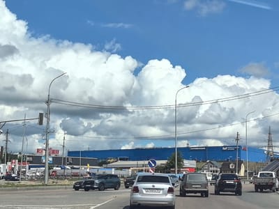 The image captures a view from a vehicle on a multi-lane road in Зоринский, Russia, on a cloudy day. In the foreground, several cars are driving on the asphalt road, including a silver Volkswagen sedan with license plate A744PA 163, a dark SUV, and a white pickup truck. The middle ground features a street with more vehicles and buildings. Prominently visible is a large, blue building with "СТРОЙЛАНДИЯ" and "Hoff" written on it, suggesting a retail or commercial complex. Signs for "МЕБЕЛЬ МОЛЛ" and "BOSS" are also present. In the background, a vast expanse of blue sky is filled with large, white cumulus clouds. Power lines and streetlights are visible throughout the scene, indicating an urban or suburban setting.