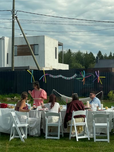In Simagino, Russia, a group of people are gathered for an outdoor celebration, likely a birthday, as indicated by a banner that appears to read "С ДНЕМ РОЖДЕНИЯ" (Happy Birthday). The scene is set in a backyard with a lush green lawn and a dark wooden fence. A white tablecloth covers a long table laden with food and drinks, with white folding chairs arranged around it. Several individuals are seated at the table, engaged in conversation and enjoying the event. Two people stand, one in a red and white striped dress, holding a plate. Colorful balloons, some arranged in starburst patterns, are attached to the fence, adding a festive touch to the atmosphere. In the background, modern white houses and tall green trees frame the scene under a cloudy sky. A hammock hangs near the fence, suggesting a relaxed ambiance. A telephone pole with overhead wires stands prominently in the foreground, partially obscuring the view of one of the houses.