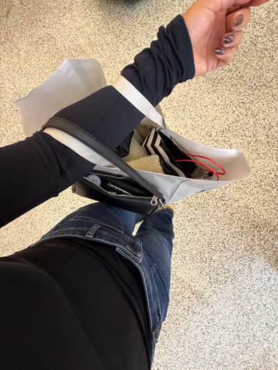 An overhead, angled image captures a person standing on a speckled, light-colored floor, consistent with an indoor commercial setting, potentially a shopping mall, in Saint-Laurent-du-Var, France. The individual wears blue denim jeans and a dark, presumably black, long-sleeved top, layered beneath another dark long-sleeved garment on the left arm. This outer garment features a thumbhole cuff and a prominent white stripe on the upper arm. The left arm carries a white paper shopping bag containing a black and white striped item, a red cord, and a cream-colored, textured material resembling faux fur or fleece. A black shoulder bag with a silver-toned zipper is also carried on the left arm. The visible left hand displays manicured nails with a dark, possibly iridescent or glittery, polish.