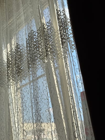 The image depicts a close-up view of a sheer white curtain with an embroidered pattern of swirling vines and circular motifs. Sunlight streams through the curtain, creating dappled light and shadows on its surface. Through the translucent fabric, a window can be seen, revealing a bright blue sky with some scattered clouds. The hint images indicate that this scene is likely indoors, looking out a window. Based on the provided information, this scene is located in the city of Kamensk-Uralsky, Russia.
