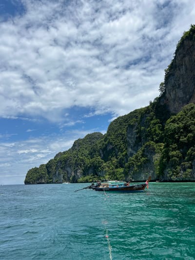 This is a photograph taken from a boat on turquoise water, looking towards a densely forested, rocky island. The water is slightly choppy, with ripples and reflections. In the foreground, a longtail boat, a traditional Thai vessel, is anchored. The boat is wooden and appears to have passengers and some cargo. It is adorned with a colorful ribbon tied to its bow. In the mid-ground, other boats are visible, and further in the distance, the imposing, green-covered cliffs of the island rise steeply from the sea. The sky above is a mix of blue sky and scattered white clouds. The image conveys a sense of tropical beauty and serene maritime activity, likely in the area of Ko Phi Phi, Thailand, known for its dramatic limestone karsts and clear waters.