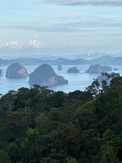 The image captures a panoramic view of a tropical seascape from an elevated vantage point overlooking a dense, verdant forest. The foreground is dominated by a thick canopy of dark green trees, with some lighter green foliage interspersed. The trees appear lush and healthy, typical of a rainforest environment.

Beyond the forest, a calm, blue body of water stretches out, dotted with numerous islands. These islands are characterized by their steep, rocky formations covered in vegetation, creating a dramatic silhouette against the water. The islands vary in size and shape, with some appearing as larger, more prominent landmasses and others as smaller, more distant formations.

In the background, a series of hazy, layered mountains recede into the distance, their forms softened by atmospheric perspective. The sky above is a pale blue, with scattered, wispy clouds, suggesting either early morning or late afternoon light. The overall impression is one of natural beauty and tranquility, characteristic of coastal Thailand.

The specific location of Ban Din Daeng Nui, Thailand, suggests this scene is likely within or near the Phang Nga Bay region, known for its striking limestone karsts and emerald waters. The image does not contain any people or human activity, focusing solely on the natural landscape.