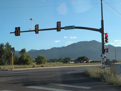 An exterior medium shot captures a street intersection in an undetermined location, United States, under a clear blue sky. A traffic light hangs over the road, with red lights illuminated on multiple signals. A street sign identifies the intersection as "Cherryvale Rd." To the left, a white and black directional sign indicates a lane change. The road is asphalt, with visible lane markings and a pedestrian crossing. In the background, lush green trees line a grassy area, and a mountain range is silhouetted against the horizon. The scene appears to be at dusk or dawn, given the lighting conditions.