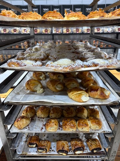 A metal rack, likely located in a bakery, holds four trays of baked goods in a commercial kitchen setting. Each tray contains various pastries. The top tray holds larger pastries with a light dusting of powdered sugar, while the second tray contains pastries with a more substantial amount of powdered sugar. The third tray holds a selection of what appear to be filled pastries, with visible fillings. The bottom tray contains pastries with a darker color and a glaze or sauce. The pastries on the top tray are likely some type of croissant. Those on the second tray appear to be similar but are likely filled. The pastries on the bottom two trays are unidentified, but the bottom tray appears to be filled pastries. The pastries are likely being prepared for sale or display. The rack is located at 31.865° N, 34.815° E, which places it in the city of Kiryat Ekron, Israel.