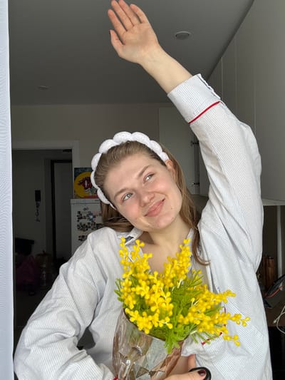 A single female subject is depicted in an indoor residential setting within Nikol'skii, Russia. The subject has light brown hair, light-colored eyes, and fair skin, exhibiting a slight upward gaze and a gentle smile. She is wearing a white, scalloped headband and a light blue and white striped long-sleeved shirt with a red trim detail on the right sleeve cuff. Her right arm is raised above her head with an open palm. In her left hand, she holds a clear, faceted glass pitcher containing a bouquet of bright yellow flowers with green foliage, consistent with mimosa. The background features a white window frame with a sheer white curtain on the left. To the right, white kitchen cabinetry with dark handles is visible. Further in the background, a refrigerator adorned with magnets and a circular colorful artwork, possibly depicting a bovine head, are discernible.