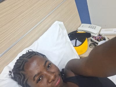 This indoor scene, located in the city of Close, Nigeria, features an individual lying on a white pillow, likely on a bed. The person is a young Black female with dark skin, braided hair styled in cornrows with wavy edges near the temples, and a slight sheen on her forehead. She is looking directly at the camera with a subtle smile. Her right arm is visible in the foreground, extending towards the viewer, indicating a self-portrait. She is wearing a dark, possibly black, top.
To the right of the individual's head, on a light wood-grain surface serving as a bedside table or nightstand, a white corded landline telephone with a push-button keypad is positioned. Next to the telephone are several colorful beaded accessories and black wires. A black baseball cap with a yellow brim and a white "A"-shaped or triangular emblem is placed upside down on the nightstand. The background consists of light wood-grain paneling with a thin, silver-colored horizontal strip, and a section of a blue accent wall is visible in the upper right.