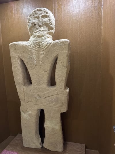 This is a full-body, eye-level shot of an ancient anthropomorphic stele, viewed from the rear. The stele is carved from light-colored stone and depicts a standing, headless figure with a stylized beard. The figure has a wide torso with openings that suggest arms, and a narrow lower body split into two legs. Intricate carvings adorn the stele, including concentric circles around the neck and geometric patterns on the waist. The stele stands on a textured platform, possibly wood or stone, and is positioned against a vertically textured, brown wall. To the left of the stele, a small portion of a informational placard is visible, containing text in an unidentified script. The overall environment suggests a museum or exhibition space.