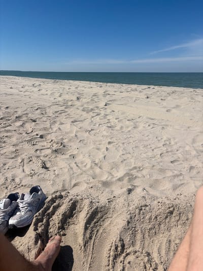 In the city of Povarovka, Russia, a pair of gray athletic shoes rests on a sandy beach. The shoes are positioned on a small sand dune, with the toes of a bare foot visible in the foreground to the right. The sandy beach extends towards the horizon, where it meets the calm, blue water of the sea. The sky above is a clear, bright blue with a few wispy clouds. The overall scene depicts a relaxed moment on a sunny day at the seaside.