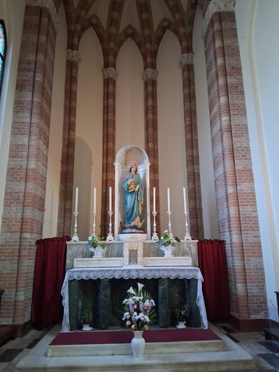 This is a vertical, eye-level interior shot of a church altar. The altar features a statue of the Virgin Mary holding the infant Jesus, dressed in blue and gold robes, set within a niche. To the right of the statue, a white lace-covered altar display holds floral arrangements and religious artifacts. Three white candles on ornate silver stands are placed in front of the altar. The scene is framed by red velvet curtains and brick archways with white accents, suggesting an older, traditional church interior. The lighting is soft and diffused, indicating an indoor setting, possibly during daylight hours. No text is visible in the image. The overall atmosphere is serene and reverent, consistent with a place of worship.