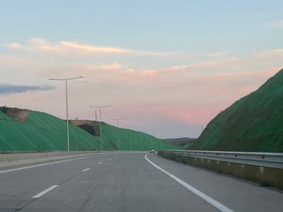 The image depicts a multi-lane highway traversing a landscape in Akhalsopeli, Georgia, during either sunrise or sunset, indicated by the light blue sky transitioning to soft pink and orange hues near the horizon with scattered clouds. The road, composed of dark asphalt with distinct white lane markings, appears well-maintained. From a low perspective, likely from a vehicle, the road curves gently to the right as it recedes into the distance.
On both sides of the highway, steep embankments or hills rise, largely covered with a vibrant green, textured fabric or netting, possibly for erosion control. Some areas, particularly on the left hill, expose patches of brown earth. A series of modern, T-shaped streetlights with unlit fixtures line both sides of the road, extending into the background.
To the left, a concrete median barrier separates the lanes. To the right, a silver metal guardrail with red reflector dots runs along the roadside, and reflections are visible on the surface below it. In the distance, a single white vehicle is observed traveling ahead in the rightmost lane. No people are visible.