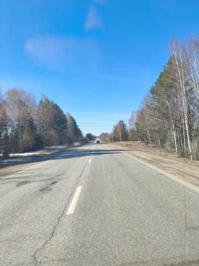 A medium shot captures a straight, paved road stretching into the distance under a clear blue sky. On either side of the road, dense forests with a mix of evergreen and deciduous trees flank the asphalt. To the left, patches of snow linger near the tree line. A single vehicle is visible in the distance on the road. Road markings include a single, solid white line indicating lane separation. The road surface shows signs of wear, with cracks and patches. The location is in the vicinity of Kukui, Russia, as per the prompt.