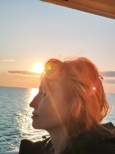 A young woman with short, reddish-blond hair is captured in a profile view, looking out towards a body of water. She is positioned in the foreground, with the sun setting behind her, casting a warm glow and creating lens flares across the image. The sun is low on the horizon, its light reflecting brilliantly on the rippling water. A faint horizon line separates the sea from the pale orange and blue sky, with a few wispy clouds present. The woman is wearing a dark garment. The setting appears to be an outdoor location, possibly a boat or a pier, given the overhead structure visible in the upper portion of the frame. The scene evokes a sense of tranquility and contemplation. The location in Kakkarovo, Russia is inferred from the provided context but is not visually identifiable from the image itself.