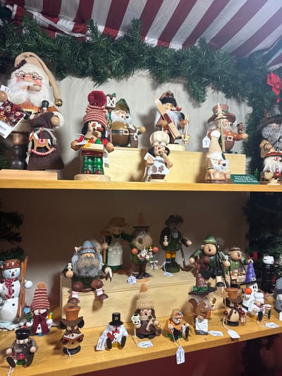 Two shelves display wooden Christmas ornaments, divided by a vertical wooden panel. The left shelf features ornaments of Santa Claus, skiers, and chefs, set against a backdrop of a red-and-white striped curtain and green garland. The right shelf contains ornaments depicting hunters, snowmen, and various characters in traditional attire. Price tags dangle from several ornaments. The scene suggests a Christmas market or holiday display.