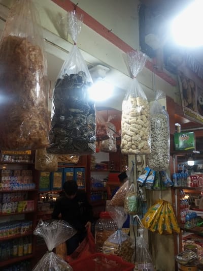 The image depicts the interior of a retail establishment, likely a snack shop or market stall, located in Lê Thanh Nghị Ward, Vietnam.
In the foreground and midground, numerous large, clear plastic bags filled with various packaged snacks are suspended from an overhead structure, tied with red ties and secured with nails. From left to right, these include a bag of brown, irregular, crispy items; a bag of dark, spiraled, pattern-embossed cookies or crackers; a bag of light-colored, sesame-coated, elongated snacks; and a bag of white, irregularly shaped items. Below these, smaller bags of snacks are visible.
The background features wooden shelves stocked with a dense array of packaged goods, including boxes, bottles, and smaller bags of various products, suggesting groceries and beverages. A dark-haired individual, wearing a dark long-sleeved shirt or jacket, is positioned in the lower-mid section, bent forward, appearing to handle items within a red plastic basket or bag. Their face is obscured. To their right, partially visible, is another individual wearing a dark cap or head covering and a blue garment.
Above the shelves on the right, a rectangular sign with faded white text, including "TOKO KUENA," is affixed to a wooden beam. Artificial lighting, consisting of bright overhead LED or fluorescent bulbs, illuminates the scene, creating reflections on the plastic bags. Smaller packets and sachets of snacks in blue and yellow packaging also hang from vertical supports on the right side of the frame. A large clear plastic jar with a red lid, containing snacks, rests on a surface in the lower right corner. The environment appears functional and densely packed with merchandise.