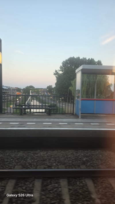 This is an eye-level, medium shot of a railway track and platform in Nieuwerkerk aan den IJssel, Netherlands. In the foreground, the dark-grey ballast of the railway track is visible. The platform edge, marked with white lines, separates the track from the platform. Beyond the platform, a metal fence encloses a station area. To the left of the fence, a black metal bench sits on the platform. To the right of the fence, a modern bus shelter with a grey roof and blue and red panels is visible. In the background, a canal runs through the city, with buildings and trees lining its banks. The sky is a pale blue with a faint glow of sunset. The image has a watermark at the bottom left that reads "Galaxy S26 Ultra".