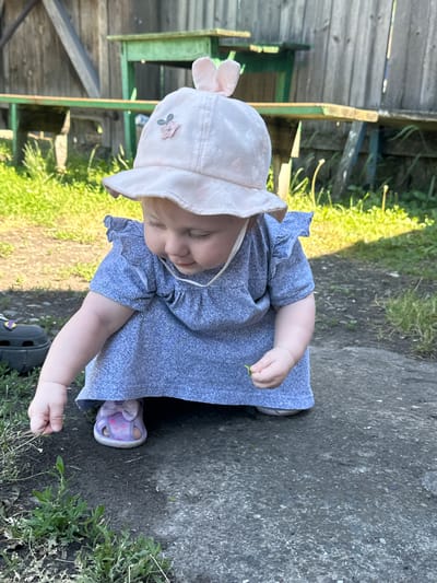 The image shows a young child, likely a toddler, kneeling on a dirt and gravel surface in what appears to be a backyard. The child is wearing a light pink sun hat with bunny ear accents and a light blue textured dress with ruffled sleeves. They are reaching down to pick up a small green item, possibly a leaf or a piece of grass. In the background, there is a weathered wooden fence and a green wooden structure, suggesting a rural or suburban outdoor setting. The lighting indicates it is daytime, with sunlight casting shadows on the ground. The overall impression is of a child exploring their surroundings in a natural environment. The location is identified as Novoyeniseyka, Russia.