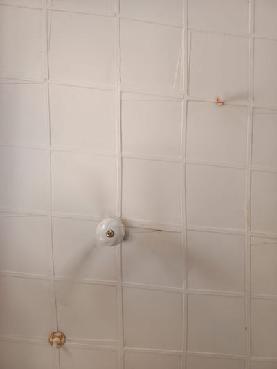 The image displays a detailed, upward-facing view of a white paneled ceiling located in Anguwar Jaba, Nigeria. The ceiling is constructed from numerous off-white, slightly textured square or rectangular panels, arranged in a grid pattern. The seams between the panels are demarcated by raised, painted lines, exhibiting minor irregularities and evidence of wear, including subtle cracks and minor discoloration.
A three-bladed white ceiling fan, with a central gold-colored cap, is centrally positioned in the lower middle section of the frame and appears to be in motion, indicated by the blurred appearance of its blades.
In the upper right quadrant of the ceiling, a bare, frosted white light bulb is affixed directly to a panel via a visible orange-colored socket. In the lower left quadrant, a second light fixture is present. This fixture features a smaller, clear light bulb emerging from an ornate, circular base decorated with gold-colored scrollwork patterns.
No individuals, discernible actions, or interactions are present within the frame, which solely focuses on the architectural elements and fixtures of the ceiling.
