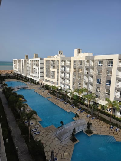 This image depicts a resort or hotel complex in Al Ahia', Egypt, featuring multiple multi-story buildings with a mix of stone and white facades, balconies, and visible air conditioning units. A series of connected swimming pools runs along the length of the buildings, edged with stone walkways, lounge chairs, palm trees, and other greenery. A small white bridge spans one section of the pool. In the distance, the Red Sea and a beach with sun umbrellas are visible. The sky is clear and blue. There are no people directly visible in the image.