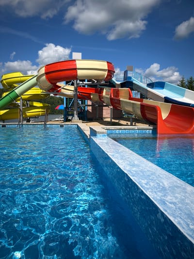 The image depicts a water park on a sunny day, with bright blue skies and white, fluffy clouds overhead. In the foreground, a section of a shimmering blue swimming pool is visible, with sunlight creating intricate patterns on its surface. A tiled edge of the pool forms a clean line, leading the eye towards the main attraction: a complex of colorful water slides.

The slides are a dominant feature, showcasing vibrant hues of red, yellow, green, and blue. Some are enclosed tubes, while others are open flumes. They are supported by a metal framework, suggesting a multi-level structure. In the background, beyond the slides, a line of trees can be seen, hinting at a natural setting. A few lounge chairs and a small red and blue umbrella are scattered around the base of the slides, implying a place for relaxation.

There are no people visible in the image. The scene is static and focuses on the architecture and environment of the water park. The overall impression is one of a recreational facility, ready for visitors on a warm day. The location is identified as the city of Юбилейный, Russia.
