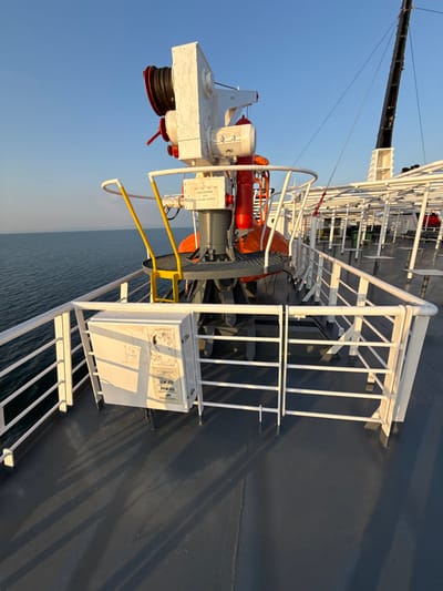 The image displays a clear, sunlit view from the port side deck of a ferry at sea, located in the vicinity of Kato Karioutes, Greece. The central element is a prominent white and grey fire monitor (water cannon) mounted on a grey pedestal. This apparatus features a black hose reel on its left side, a red cylindrical component to its right, and yellow U-shaped handles at its base. Directly below the monitor, an orange ring-shaped object, possibly a life buoy, is visible.
In the foreground and extending along the left edge, a white metal safety railing separates the grey deck from the dark blue, calm sea. A rectangular, weathered white control box, adorned with diagrams and partially legible text, is affixed to the railing structure in the lower-left corner. Elongated shadows from the railings and the central equipment are cast across the grey deck, indicating direct sunlight from the right.
Towards the midground and right, additional white railings and structures are visible, along with glimpses of empty white tables and chairs, suggesting a seating area. A tall, dark-colored crane or mast is discernible in the upper-right background against a clear blue sky. A subtle landmass is visible on the distant horizon to the left. No individuals are present in the scene.