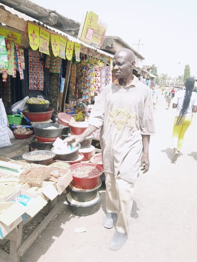 An outdoor market scene in Kadoma, Zimbabwe, is depicted under bright daylight. In the foreground, an older, bald Black male, wearing a light-colored long-sleeved shirt with a patterned chest and matching trousers, stands facing partially left with his right hand extended towards a small white plastic bag among market goods. He wears grey slip-on shoes. To his left, a wooden table displays goods in cardboard boxes, including smaller packaged items. Behind this, various dry goods like grains and legumes are visible in stacked red and black plastic bowls. A green plastic basket also holds items. Behind these displays, numerous packaged goods hang from a stall structure, above which yellow banners with red "Maggi" branding are strung. A larger "Maggi" sign is on the stall roof. The stall itself appears constructed from brick or mud brick with a corrugated roof. In the midground, a dirt path extends into the distance, where a slender individual in a white t-shirt and bright yellow leggings with long dark braids walks away from the viewer. Other indistinct figures are visible further down the path, bordered by sparse vegetation and distant structures.
