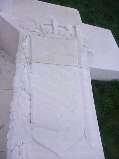 A stone structure features a central blank shield shape, likely for inscription. Above, a carved sword rests horizontally. The shield is framed by detailed carvings of leaves on both sides, suggesting oak or laurel. The stone is light-colored and weathered, set against a background of grass, possibly in a cemetery or memorial garden. The overall impression is one of solemn remembrance.