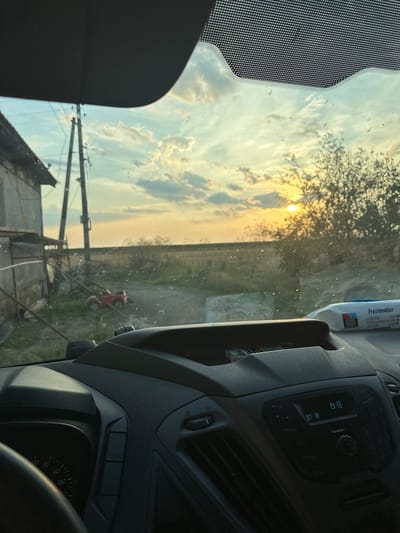 The image, taken from inside a vehicle, captures a sunset scene in Mlekarevo, Bulgaria. The view is framed by the car's interior, including the dashboard and windshield. The windshield is speckled with dirt or water droplets. Outside, a rural landscape unfolds with a house and a utility pole on the left, a dirt road leading into the distance, and trees on the right. A small red toy car is visible on the dirt road. The sun is setting, casting a warm glow through the trees and clouds in the sky. On the dashboard, a packet of "Freshmaker" wet wipes is placed next to a cigarette pack. The car's radio display shows the time as 19:19.