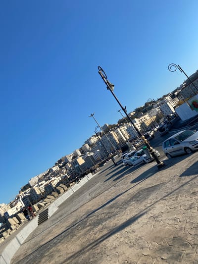 The image shows a street view in Beau Fraisier, Algeria on a clear day. The camera is pointed upward at an angle, capturing the clear blue sky, the facades of buildings, street lamps, parked cars, and a textured sidewalk. The buildings appear to be densely packed and of medium height, with a variety of architectural styles. Street lamps with ornate curved arms are visible along the street. Several white cars are parked parallel to the sidewalk. Shadows cast by the lamps and buildings indicate bright sunlight. The sidewalk has a cracked, textured surface. Some unidentified figures can be seen along the street.