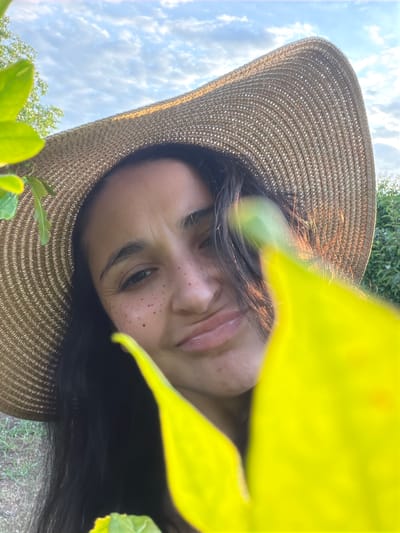 The image is a close-up, selfie-style shot of a young woman wearing a wide-brimmed straw hat. She has dark hair and is looking towards the camera with a slight pout. Her face is adorned with freckles, and she has wrinkles around her eyes. The foreground is blurred by bright yellow leaves, and the background shows a sky with white clouds and green foliage. The location is identified as Ароматненское сельское поселение, Ukraine, suggesting an outdoor, possibly rural setting.