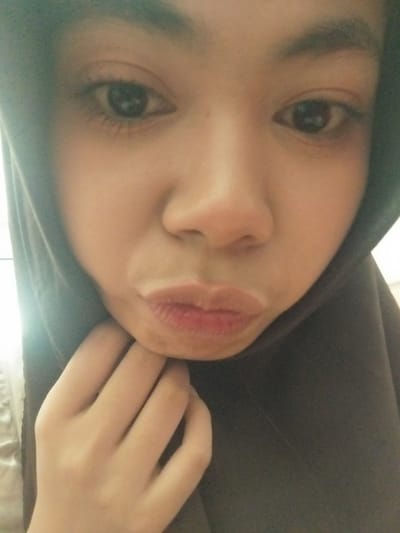 The image is a close-up selfie of a person wearing a dark hijab. The person is pouting their lips, and their right hand is raised to their chin, with fingers slightly curled. The background is dark and appears to be indoors, with some subtle lighting from the left. The person's eyes are visible, and they are looking towards the camera. The overall tone of the image is casual and personal. The location in Tibojong, Indonesia, is not discernible from the visual information provided in the image itself.