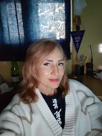 A woman with shoulder-length, light brown hair and visible makeup is seated indoors, likely in an office or room. She is wearing a white cardigan with a textured pattern over a black t-shirt with white lettering. Behind her, a dark blue curtain with a raised pattern is visible, with light filtering through it, creating shadows. To her right, a triangular blue pennant with a white logo and text hangs on the wall. On a desk behind her, a cup filled with pens and a black radio device are present. A green bottle is on a surface to her left. The overall scene suggests a personal workspace.