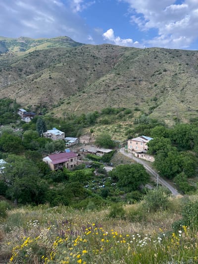 A village nestles into a mountainside, interspersed with trees and meadows. Buildings with red and blue roofs are dotted among the greenery. A road winds through the scene. Lush green mountains rise beside the village, with a blue sky and fluffy white clouds overhead. Yellow wildflowers bloom along a bank in the foreground.