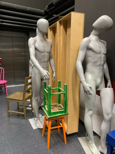 The image depicts an interior scene in Nowy Targ, Poland. The environment features a dark, possibly charcoal-grey, floor and dark walls. On the left, silver metallic elevator doors are visible, displaying a red indicator light. Above the elevator, black corrugated pipes run horizontally across the ceiling.
Two grey, male display mannequins are positioned in the foreground/midground. The mannequin on the left stands erect on a white square base, its head featureless and facing slightly to the viewer's right. Its right arm is bent, with the hand extended forward. A second grey mannequin is partially obscured behind the first. The mannequin on the right is also grey, standing on a white square base, but its head is tilted back and to the left, partially obscured by a light-brown wooden partition. Its right lower leg, from the thigh down, is detached and stands upright adjacent to its left leg.
Various chairs and stools are arranged within the scene. A pink metal chair with a curved back is partially visible on the far left, near the elevator. A golden-colored wooden chair is positioned to the left of the leftmost mannequin. In the center foreground, an orange four-legged wooden stool is upright, with a green four-legged wooden stool inverted and resting on top of it. The green stool's top surface (now downward-facing) shows exposed wood. A blue object, possibly a stool or crate, is partially visible on the far right of the frame, near the detached mannequin leg. A tall, light-brown wooden partition or shelving unit stands vertically between the two main mannequins, extending towards the ceiling.