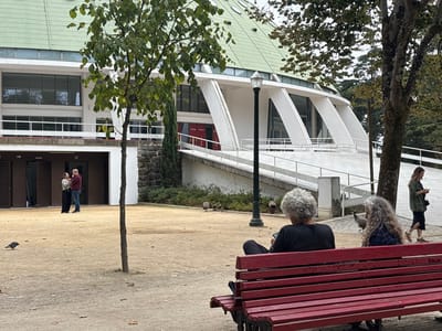 The image depicts an outdoor scene within the Jardins do Palácio de Cristal, featuring the Pavilhão Rosa Mota (Super Bock Arena), located in Lordelo do Ouro e Massarelos, Porto, Portugal, under an overcast sky.
In the immediate foreground, two individuals with grey, curly hair are seated from behind on a red slatted park bench. The individual on the left, wearing a dark top, holds a mobile phone. The individual on the right, with longer grey hair and a patterned dark top, also holds a mobile phone. A pigeon stands on the ground near the left side of the bench.
The midground features a large, light brown dirt area, with a slender tree centrally positioned. Another pigeon is present on the ground to the left of the tree. A tall black lamppost with a white fixture stands between the bench and the main building. To the left in the midground, a man in a red long-sleeved shirt and blue jeans stands facing a woman in a light top and dark skirt; they appear to be conversing in front of multiple dark brown, shuttered openings on a lower level of a building.
The background is dominated by the Pavilhão Rosa Mota, a large structure with a prominent green geodesic dome roof and a white facade featuring bands of horizontal windows. A wide, white concrete ramp, supported by angular white pillars, sweeps upwards towards the building's entrance. On the right side of this ramp, a woman with light hair, wearing a dark green jacket and a dark skirt, walks while looking at a mobile phone. Additional trees and general foliage are visible in the far background to the right.