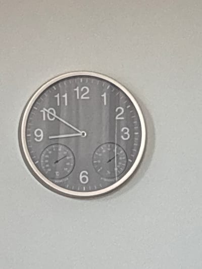 A round, grey wall clock with white numbers and hour markers is mounted on a light grey textured wall. The clock has two smaller circular dials inset at the bottom, one on the left and one on the right. The clock hands indicate approximately 9:50. The clock is the sole object in the frame. There is no identifiable location in the city of Cork, Ireland.