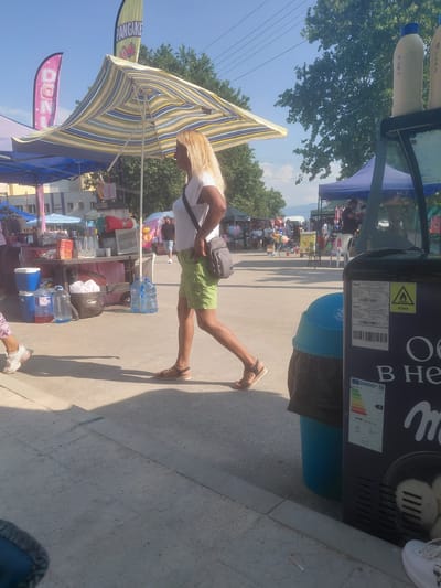 The image depicts an outdoor market or fair taking place in Kostievo, Bulgaria, under a clear blue sky. The ground consists of a paved surface, likely concrete or asphalt, showing some wear. In the foreground, slightly right of center, a woman with long, straight blonde hair walks from left to right in profile. She wears a white short-sleeved t-shirt, olive green shorts, and brown flat sandals. A grey messenger bag is slung over her left shoulder. To her immediate left, a child's left foot and lower leg are partially visible, wearing white sneakers with pink accents and light-colored patterned pants, also moving from left to right. Various market stalls and tents populate the scene. On the left, a blue-canopied tent covers a table laden with merchandise, including plastic cups, red packages, a red beverage dispenser, a silver appliance, and several large plastic water bottles. A blue cooler with a white lid and a black trash bag containing white items rest on the ground near this stall. Behind this stall, a light-colored building with windows is visible. Two prominent feather flags are displayed: a pink one on the far left reads "DONUTS" with a donut graphic, and a greenish-yellow one behind a large, open, striped (yellow, green, blue, white) market umbrella reads "PANCAKE" with fruit graphics. On the far right, a tall, dark-colored commercial refrigeration unit is visible. On its top, two cream-colored cylindrical containers with blue caps are present, marked with Cyrillic script: "КУБИ" (CUBES) and "ЯГОДА" (STRAWBERRY). The unit displays a yellow warning triangle, an "R290" refrigerant label, a barcode, and an energy efficiency label indicating "ENERGY D" and "949 kWh/annum". Inside the unit, white, round items resembling ice cream or frozen yogurt are visible. Adjacent to the refrigeration unit is a blue trash bin with a black liner. In the mid-ground and background, additional market tents and numerous indistinct people are present. One man in a dark shirt and light shorts is visible standing behind a blue tent further back on the right. Another man in a dark shirt is partially obscured behind the left stalls. Further down the street, various market goods like colorful balloons, pinwheels, and what appears to be cotton candy are visible. A white plastic chair is situated near one of the background stalls. The background features abundant green trees against the clear blue sky, with overhead power lines traversing the upper portion of