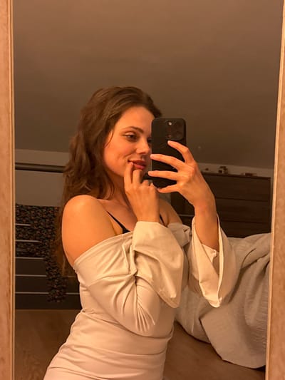An image captured as a mirror selfie displays a female subject indoors in Cava de' Tirreni, Italy. The subject, possessing long, wavy brown hair, is positioned from the chest up, angled to her left. She is attired in a light-colored, off-the-shoulder top or dress, with a black bra strap visible on her left shoulder. Her left hand holds a dark-colored smartphone, angled to take the photograph. Her right hand is placed near her mouth, with fingertips touching her lower lip. Her expression features a subtle smile, and her gaze is directed towards the left. The reflected background reveals an interior space with warm ambient lighting. Visible elements include a clothing rack displaying dark, patterned fabric on the left, and a bed with a light-colored blanket and a dark wooden dresser on the right. The mirror itself is bordered by a light-toned frame.