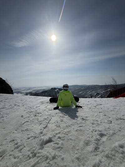 The image displays a solitary individual seated on a snow-covered mountain slope in Zelenaya Polyana, Russia, under a bright, sunlit sky. The person is viewed from behind, dressed in a neon green jacket, dark lower garments, black gloves, and a white helmet with goggles. A black snowboard is positioned on the snow directly behind the individual. Their arms are extended to the sides, with hands resting palms down on the snow. A distinct shadow of the person is cast forward onto the textured snow surface. The background features a panoramic view of snow-dusted mountain ranges and valleys under a light blue sky with faint cirrus clouds. A dark rock formation and bare tree branches are visible on the left side of the frame, while a wooden fence and a partial red object are present on the far right. A diagonal multicolored lens flare is visible in the upper right sky quadrant, originating near the sun.