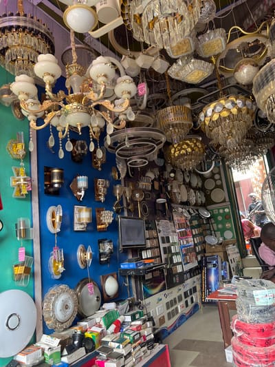 The image displays the interior of an electrical and lighting retail store in Igbede, Nigeria. The ceiling is densely packed with a wide assortment of chandeliers and suspended light fixtures, featuring various designs including ornate crystal and glass chandeliers with gold-toned metalwork, as well as modern circular and rectangular fixtures. The left wall, painted in a vibrant blue, showcases numerous wall-mounted sconces of different styles, some with attached product tags.
In the lower foreground, a red-edged counter holds stacks of small, packaged electrical items, likely switches or small components. Above this, a black flat-screen monitor is mounted on the wall. Adjacent shelves display rows of electrical switches and wall plates. In the lower-right foreground, a person wearing a light-colored shirt is seated with their back to the camera. Next to them, rolls of red and black electrical wire are stacked on a wooden table. Towards the rear of the store, an entrance area reveals a partially visible person standing, and outside, another individual is seen on a motorcycle. A large white standing fan is partially visible on the far right. The environment is characterized by a high density of merchandise, indicating a specialized commercial establishment.